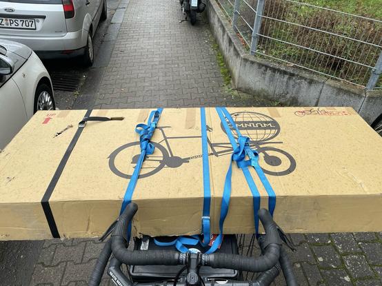 Ein großer Karton auf einem Lastenrad aus Fahrerperspektive. Der Karton ist quer uber das Rad gelegt und mit Spanngurten befestigt. Auf dem Karton ist die Silhouette eines Lastenrads und die aufschrift Omnium gedruckt