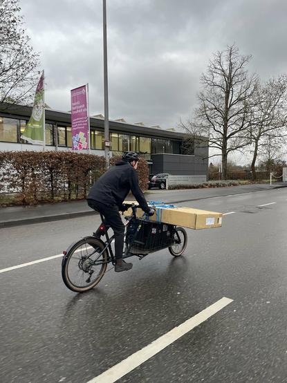 Bild einer Person, die auf der Fahrbahn mit dem Lastenrad einen großen Karton transportiert. Der Karton ist quer vorn auf das Rad geschnallt und ragt an beiden weit über.
