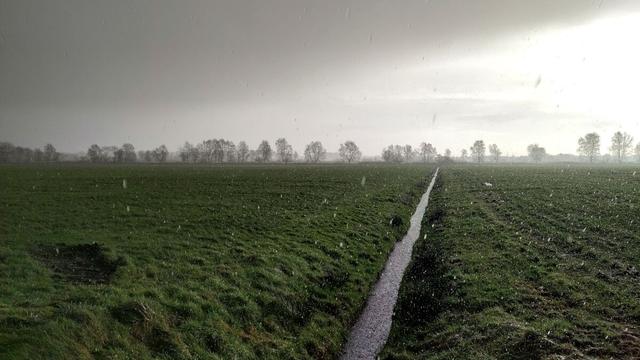 View over grassland into the distance. Slightly to the right of the center, a water-bearing ditch runs to the horizon. The water is turbulent from the falling precipitation. The floodplain is full of small, white hailstones. Some of them leave streaks on the picture due to their movement. In the distance, a row of bare deciduous trees stands across the entire picture. Above them is a gray sky, which becomes increasingly brighter to the right.