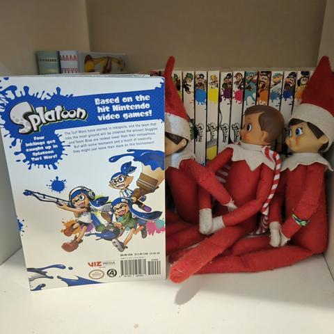 A group of three Elves on the Shelf sitting together reading Volume 1 of the Splatoon manga. They are sitting on a white bookshelf with a row of other volumes of Splatoon manga behind them. One of them, in the middle, is wearing a cape instead of a hat.