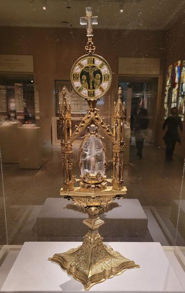 A gilded copper and silver reliquary stand about half a meter tall and with floral designs all over the square base. On the stand, at center, is an egg-shaped chunk of glass with a small cavity in its center containing a tooth. On either side of the glass are two towers supporting, above the glass, a roundel with a depiction of the crucifixion. At the very top is a small crystal cross.
