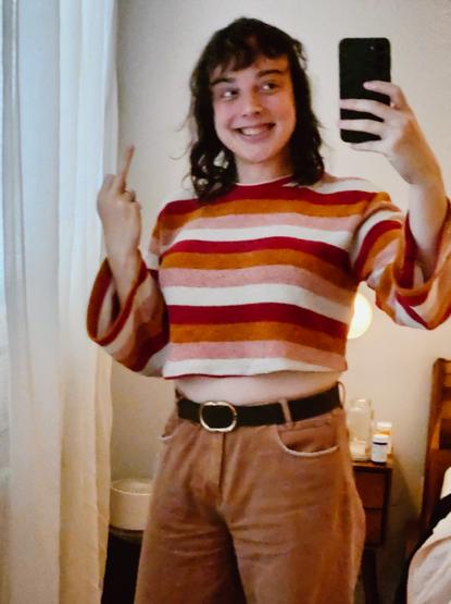 stel smiling in orange pants and a lesbian flag crop sweater. shes giving a middle finger also