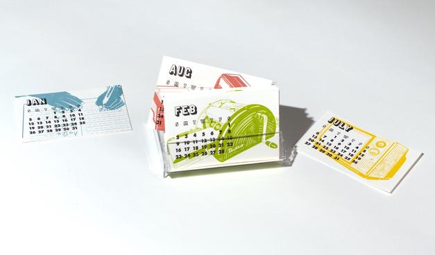 Small letterpress printed cards featuring various calendar months. The cards for February and August are in the cassette tape case. July and January lie nearby. The card for July features an old TV set in yellow. February features a mimeograph machine printed in green. Each card shows the calendar days in a grid with the weekdays.