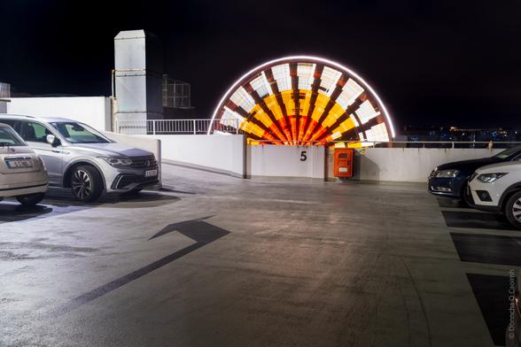 A nighttime parking garage scene with cars and a brightly lit, colourful half-circle light made by the big wheel spinning. The number "5" is visible on the wall.