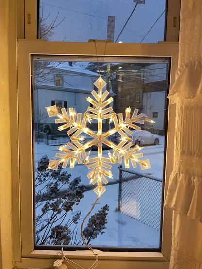 A translucent plastic snowflake, about 16" in diameter, dangling flush against a small window. The snowflake is lighted with a couple dozen small bulbs.  To the left of the window a bit of ruffled eyelet curtain is hanging. Below the lighted snowflake its electrical cord is dangling and taped to the windowsill. Through the window is a twilight scene of snow falling gently, gathering on the street and cars and houses for the first time this season.