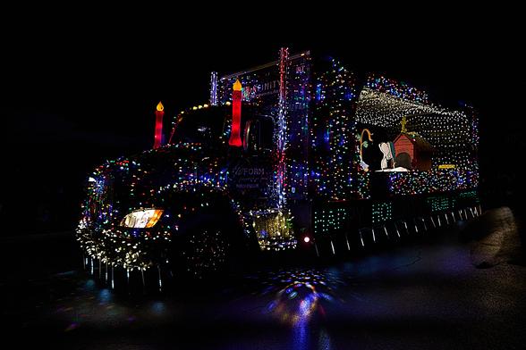 This image shows one of the trucks in the 2024 IEOA Lighted Truck parade and food drive. A soft-panel truck, it's heavily decorated with coloured lights and has two large plastic candles on either side of the windshield. The open area in the back has a scene from the Charlie Brown Christmas, featuring Charlie Brown, the small tree, Snoopy and his doghouse.