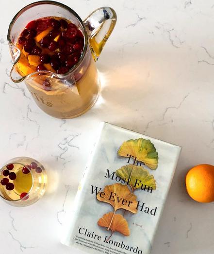 A pitcher of sangria and a glass of sangria is off to the left. The book, The Most Fun We Ever Had by Claire Lombardo is next the glass, an orange is off to the right.