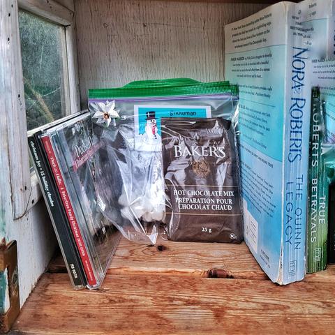 This image shows the interior of a nearby Little Library. In addition to the books on offer there are two sandwich bags, stapled together. One contains a packet of hot chocolate mix powder; the other contains ½ cup of Snowman Poop (mini-marshmallows) ⛄