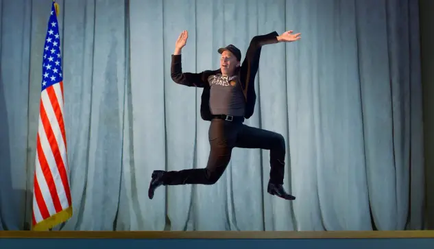 Elon Musk jumping in front of an American flag. The photo has been edited to shape his body into the form of a swastika. Credit: https://www.der-postillon.com/2024/12/musk-sprung.html