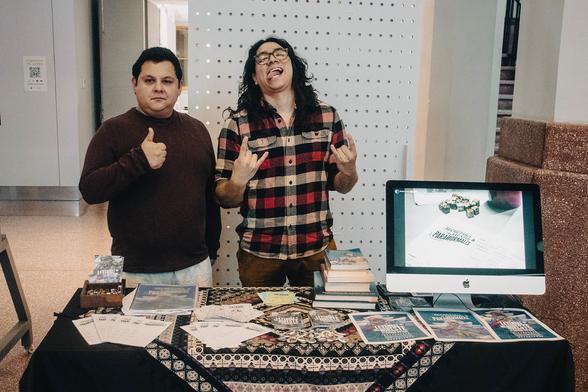 Fotografía. Dos personas detrás de una mesa con parafernalia de "Secretaría de Asuntos Paranormales", juego de rol desarrollado en el LabNL. Hay un monitor mostrando un slideshow de fotografías.