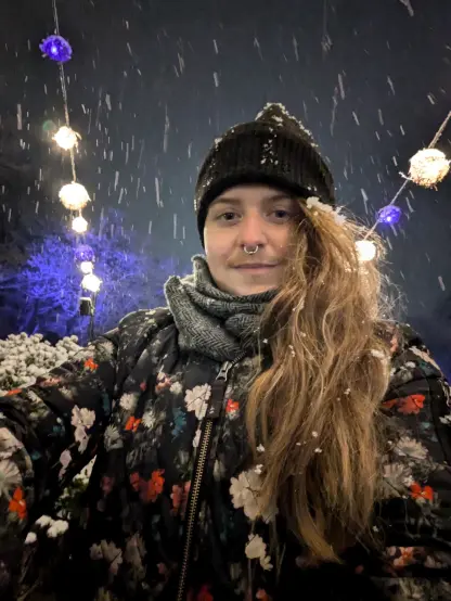 FoxFux wears a quilted floral bomber jacket with a black beanie that his redhair sticks out under as snow falls around him and settles in his long hair. There are floating ball lights in blue and white and the trees are lit up blue