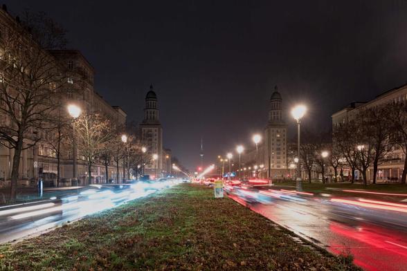 The two tower blocks of the Frankfurter Tor. A grassy strip is flanked by roads, rows of trees and street lights and buildings. In the centre is the TV tower. The cars are blurred and their lights form lines by the long exposure time.