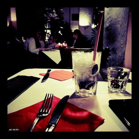 Dunkles Farbfoto, aufgenommen am Abend in einem Restaurant. Im Vordergrund die Platte eines Tischs mit Serviette, Besteck und zwei Gläsern. Im Hintergrund ein weiterer Tisch mit Gästen, dahinter die Deko des Lokals. Aufgenommen mit Xiaomi Redmit 10 mit Vignette for Android mit Cross Processing-Effekt.