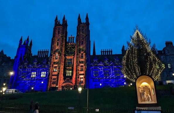 The illuminated Christmas Tree on the Mound in Edinburgh, behind a nativity scene in an illuminated shed. Behind is the New College of the University of Edinburgh, illuminated in red and blue lights with a gold pattern across it. The sky is twilight and streetlights are lit. Photo © Self