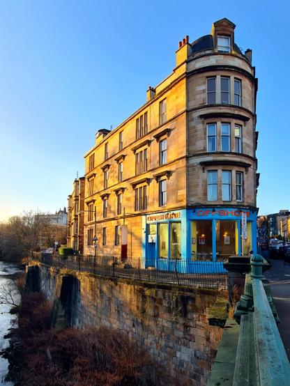 A four storey blonde sandstone corner tenement on the bank of a small river.