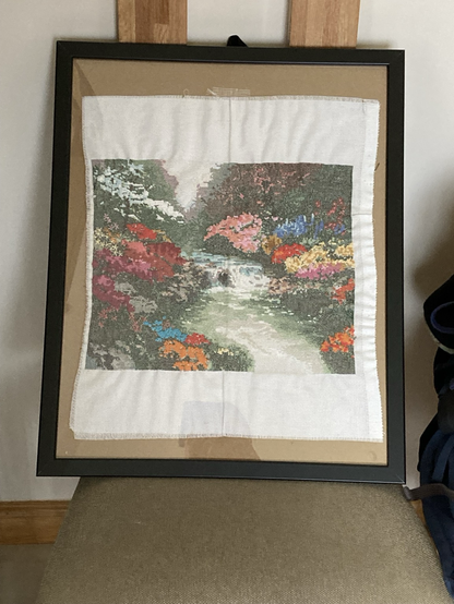 Photo of a cross stitch in a frame. Cross stitch shows a river with a small waterfall and lots of brightly coloured flowers either side with trees extending upwards.