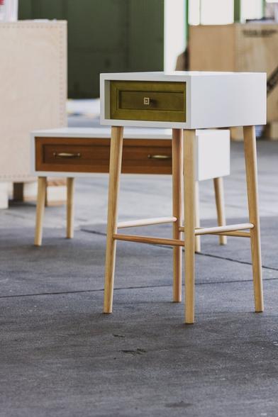 A modern side table with a white body, natural wooden legs, and a green drawer, standing on a concrete floor. In the background, another similar table with a wooden finish and brass handles is partially visible, along with industrial-style surroundings.