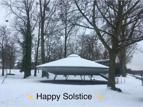 A flying saucer stands in the snow, with trees around 
Black text says ‘happy solstice’