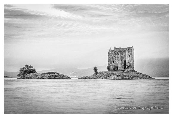 Black and white photo of a tower house on a small island in a calm sea loch. There is a smaller island to the left, and hills seen dimly in the background.
