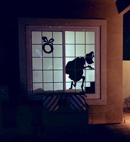 Front window of a home at night with a silhouetted Grinch that looks like he's sneaking up stairs to steal an also silhouetted Seuss-shaped wreath.