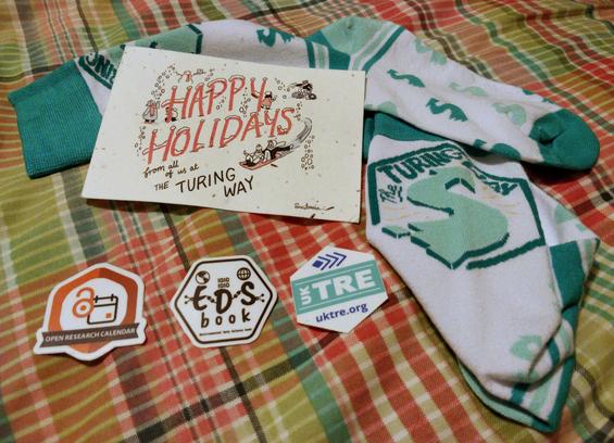 Gifts arranged on a duvet cover. There is a postcard with Christmas themed illustrations which run reads "Happy holidays from all of use at The Turing Way". Underneath are three hexagonal stickers for open research projects. At the top and right are a pair of socks patterned with The Turing Way logo and colours.