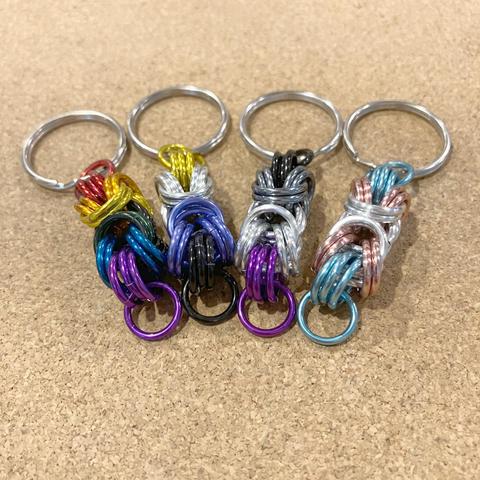 A set of four chainmail keyrings, in the colours of the rainbow pride flag, the nonbinary flag, the asexual pride flag, and the trans pride flag respectively.