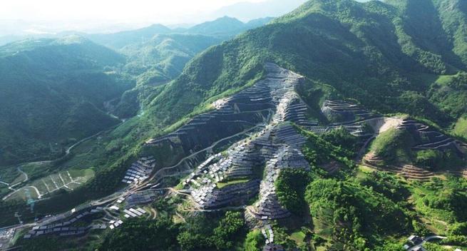Aerial photo of a hillside with many escarpments, covered in lush green vegetation, also partly covered with shiny grey solar panels. China.