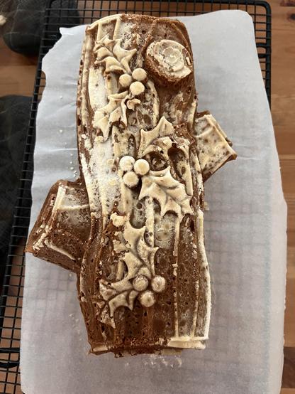 Photo of a cake shaped like a wooden log log with holly sprigs on it. The top of the cake is lighter because of the cooking spray that I had to use to ensure it turned out okay.