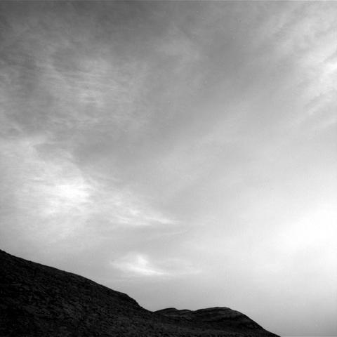 The black-and_white image shows a dark contour of hills at the bottom and lower left and a variety of whispy thin clouds in a grey sky.