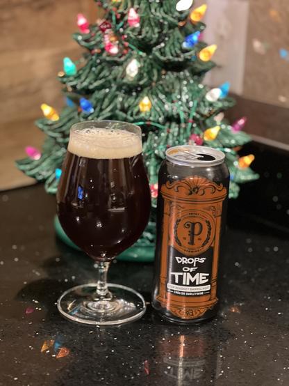 A tall tulip glass of dark beer is in front of a small lighted Christmas tree. The pint can is black with a copper colored label and lettering in white reading “Drips of Time”