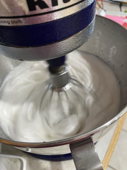 A stand mixer, whipping marshmallows into existence.
A blurry mixer whip attachment is rapidly stirring a white batter in a stainless steel mixing bowl.