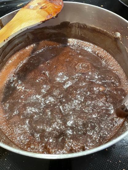 A mixture of cocoa, sugar, and milk is boiling in a stainless steel pot. A wooden spoon rests along the upper left corner of the image. A candy thermometer is barely visible in the lower right.