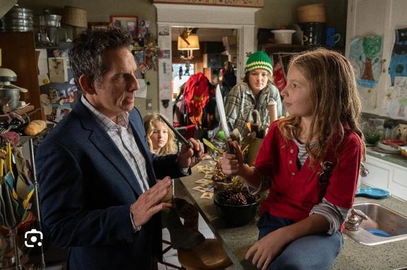 Ben Stiller is talking to a child while two other kids are watching. The  child is sitting on a kitchen counter holding a knife, and Ben Stiller is holding a wooden wand.