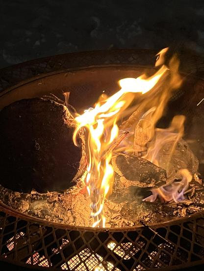 Photo looking into a fire contained in a metal fire ring. The center of the flames has a curly shape where it has been caught by the wind.