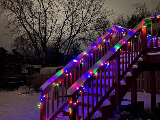 Photo of the Christmas lights on my deck railing beneath several inches of snow. The snow muffles the light and patches of it glow from within with multicolored sections.