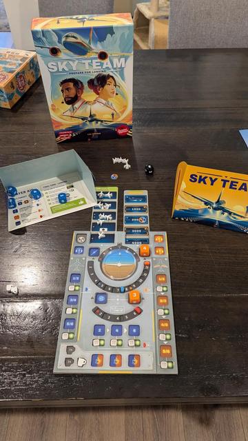 The image shows the board game Sky Team set up on a wooden table. The game box stands upright in the background, featuring colorful artwork of two pilots (a bearded man and a woman with a headset) against a backdrop of airplanes and a vibrant sky. The game components include a main game board resembling an airplane cockpit, with dice, small white airplane miniatures, and various cards and tokens. To the right, a small player screen with Sky Team artwork is positioned. The overall setup conveys a theme of aviation and teamwork.