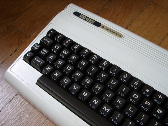 Das Bild zeigt die erste, in Japan als VIC-1001 verkaufte Version des VC-20. Ein weißer Tastaturcomputer mit einer dunkelbraunen Tastatur und einen schwarz/bronze Typenschild. Die Tasten zeigen neben Buchstaben und Commodore typischen Grafikzeichen auch japanische Symbole.