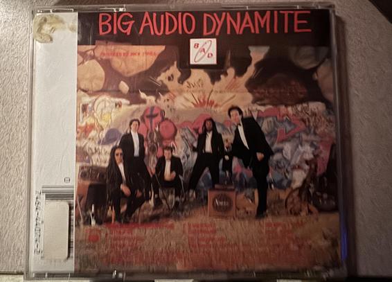 The image shows the cover of a CD by Big Audio Dynamite, featuring five band members in formal attire against a vibrant, graffiti-covered background.