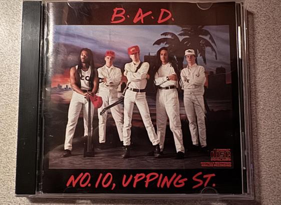 The image shows a CD cover featuring a music group named B.A.D. (Big Audio Dynamite), with five members posing confidently. They are dressed in white outfits, and some wear caps. The background includes palm trees and a cityscape, and they make me want to dress like DEVO
