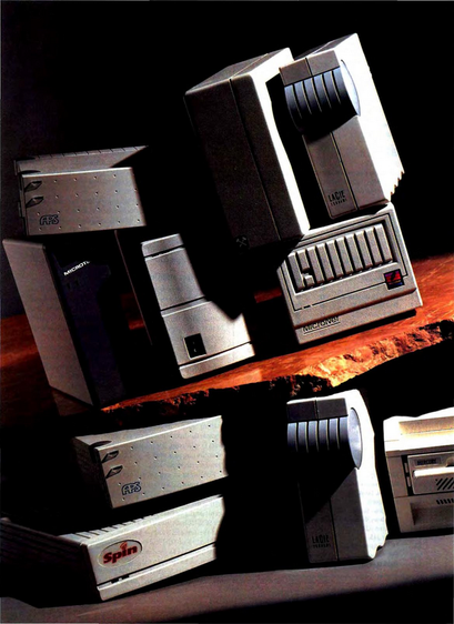 Collection of vintage external hard drives and storage devices from various brands, including APS, MicroNet, LaCie, and Spin. These devices are stacked and arranged on a wooden surface, showcasing the design and technology of retro computer peripherals