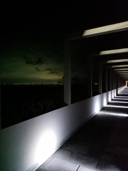 Un pasillo muy largo iluminado intermitentemente por hileras de lámparas rectangulares, al costado izquierdo la costa con luces de ciudad en el horizonte y nubes en un cielo nocturno.