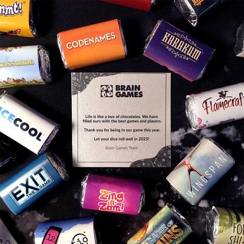 A Christmas gift from Brain Games: tiny card saying "Life is like a box of chocolates. We have filled ours with the best games and players. Thank you for being in our game this year. Let your dice roll well in 2025! Brain Games" surrounded by lots of candy wrapped in a BG board game colors + logos wrappers: Codenames, Carcassonne, Flamecraft, Karakum, Wingspan, etc