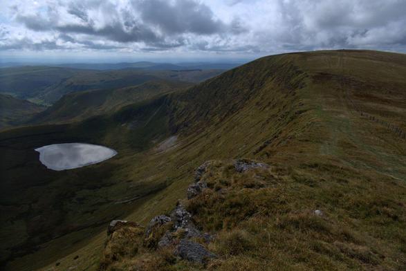 Looking along the top of a mountain escarpment. Cliffs fall steeply away on the left. A ridge leads off to the left, cradling an upland lake (Llyn Lluncaws) within its embrace. The ground rises to a summit on the right (Moel Sych) topped with a low Bronze Age cairn. It's partially cloudy and the silvery surface of the lake mirrors the cloud. Beyond the ridge on the left, hills roll away endlessly into the blue.