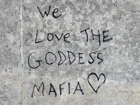 black handwritten graffiti on mottled grey stone gate pillar block reads 'We Love THE GODDESS MAFIA ❤️'