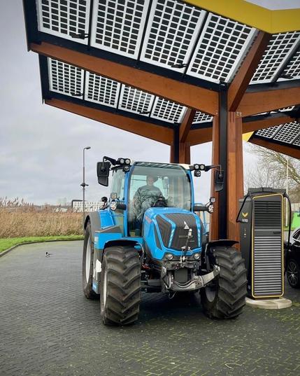 Ein blauer Traktor steht unter einem Holzpavillon mit Solarzellen auf dem Dach. Der Traktor ist modern und scheint elektrisch zu sein, da er an einer Ladestation angeschlossen ist. Im Hintergrund ist eine leicht bewölkte Landschaft zu sehen. Das Bild vermittelt den Eindruck von nachhaltiger Landwirtschaft und moderner Technologie.