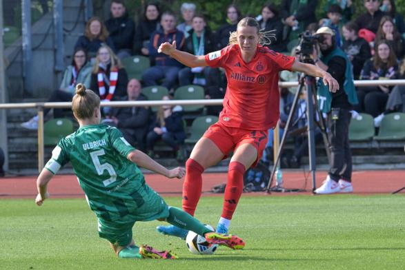 Michelle Ulbrich grätscht mit dem Rücken zur Kamera in die vor ihr angelaufen kommende Klara Bühl, die den Ball zwischen ihren Füßen hat und sich auf den Impact des Zweikampfs vorbereitet, sie hat die Arme ausgestreckt, um auf einen Fall reagieren zu können. Hinter den beiden sieht man einen Kameramann von der Fernsehübertragung und Teile des Publikums.