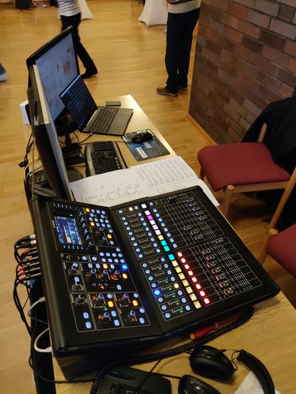 A digital audio-mixing console with lots of colourful lights, and two computer monitors and a keyboard beside it. Two chairs behind the table, shot from the side and above.