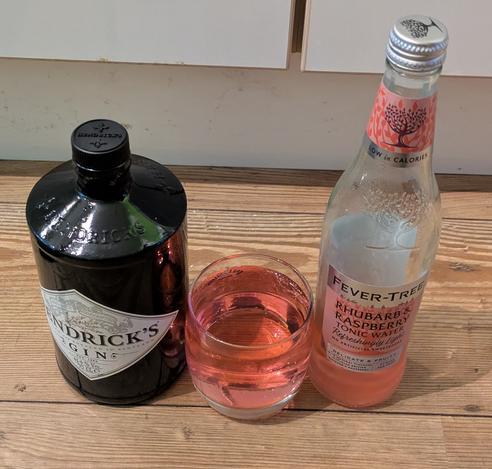 Three glass items, on the left a dark bottle of Hendricks gin, in the middle a short high ball glass with a pale pink liquid in, on the right is a clear glass bottle with Fever tree Rhubarb & Raspberry tonic on the table, there is some tonic still in the bottle.