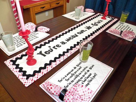 A dining room table decorated in Grinch-themed table linens. There's a table runner that reads "You're a mean one, Mr. Grinch" with the words surrounded by large black rickrack and a pink and red dot border. Four placemats, each with one of the verses from the Grinch's signature song, are placed on the table. Each placemat has a Grinch glass and a pink and red dot napkin placed artfully so you can still read the words. Finally, there are also two wonky red candle sticks with pink candles.