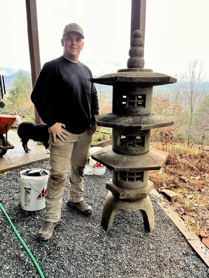 Landscaper proud of setting stone lantern on a new level base stands next to it with mini Aussie dogs wheelbarrow and bucket of stone dust nearby